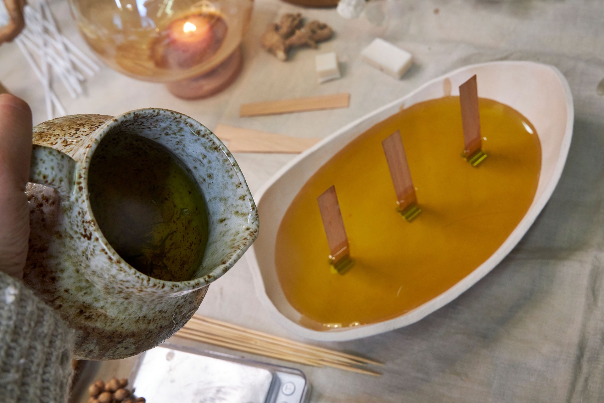 Close-up shot of the candle-making process: melted wax is being poured from a jug into a ceramic bowl with wooden wicks. A warm, handcrafted scene reflecting eco-friendly production, natural materials, and aromatherapy